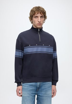 Jeune homme portant un sweat-shirt bleu marine à demi-zip avec des rayures horizontales bleu clair et le texte "HUGO", associé à un jean bleu clair.