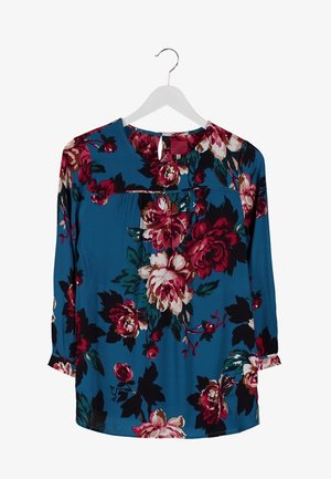 Niebieska bluzka w kwiatowy wzór z rękawami do trzech czwartę, w połączeniu z ciemnoniebieskimi skinny jeansami i czarnymi botkami, trzymająca czarną kopertówkę.