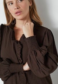 Pruun tekstureeritud blouse, millel on volangidega kaelus, volditud mansetid ja mahukad varrukad. Kanga materjal on kerge ja kortsuline välimus.