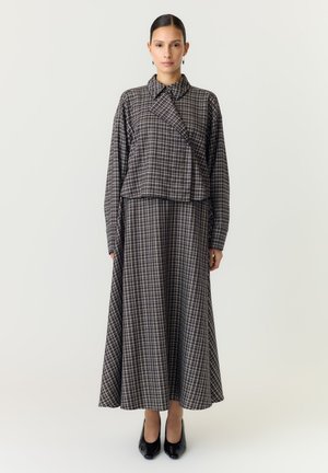 Femme debout portant une longue robe à carreaux avec une veste assortie à col et des chaussures à talons noirs sur un fond clair uni.