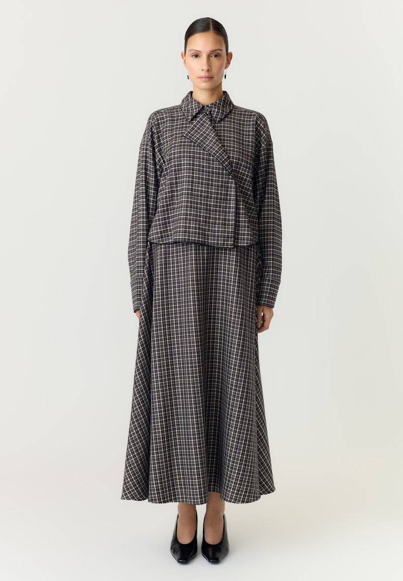Femme debout portant une longue robe à carreaux avec une veste assortie à col et des chaussures à talons noirs sur un fond clair uni.