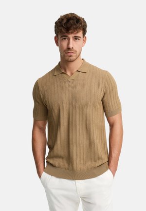 Junger Mann steht mit den Händen in den Taschen, trägt ein strukturiertes beige kurzärmeliges Strick-Poloshirt und weiße Hose, neutraler Hintergrund.