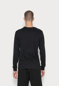 Mies, jolla on lyhyet hiukset, pukeutuneena yksinkertaiseen mustaan pitkähihaiseen paitaan ja mustiin housuihin, seisoo valkoista taustaa vasten, selkä näkyvissä.