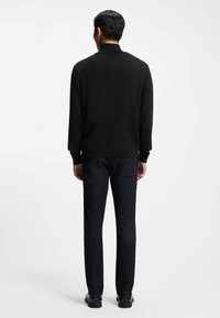 Mężczyzna ubrany w czarny sweter z golfem, ciemne dżinsy i czarne buty, stojący tyłem na jednolitym białym tle.