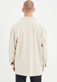 Beige oversized shirt met lange mouwen, enkele borstzak en knoopmanchetten. Gladde textuur, relaxed fit en subtiele glans.