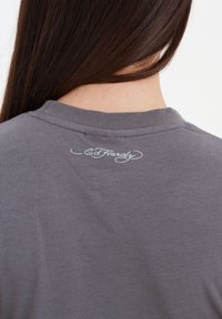 Šedé bavlněné tričko s kulatým výstřihem a malým logem "Ed Hardy" vyšitým světle modrou barvou na zadní straně límce.
