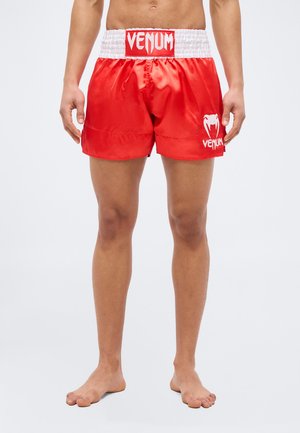 Man wearing red and white Venum branded boxing shorts, standing barefoot against a plain background.