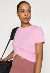 Ροζ ανοιχτόχρωμο κοντομάνικο μπλουζάκι με σχεδιασμό μπροστά που έχει στρίψιμο, κατασκευασμένο από μαλακό ύφασμα. Το μοντέλο κρατάει μια μαύρη τσάντα πάνω από κάτω μέρος σε ανοιχτό μοβ χρώμα.