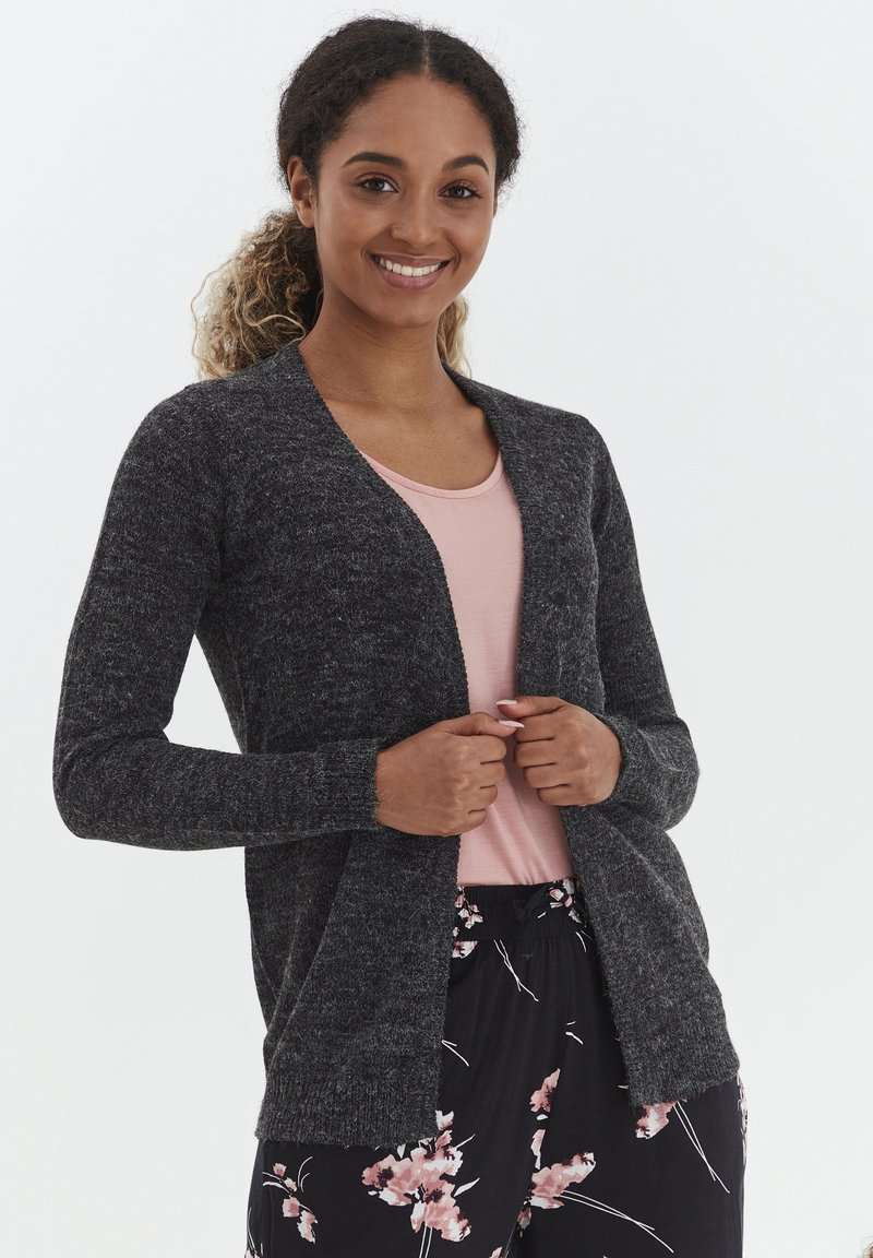 Cardigan gris foncé à manches longues, devant ouvert, en tissu tricoté texturé, porté sur un haut rose clair. Le modèle se tient devant un fond blanc.