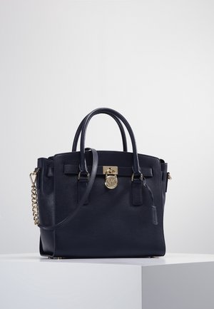 Black leather handbag with two handles, a shoulder strap, and a gold lock detail on a white pedestal against a plain background.