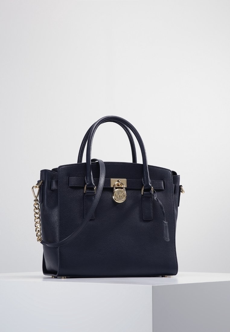 Sac à main en cuir noir avec deux poignées, une bandoulière et un détail de verrou doré sur un socle blanc sur fond uni.