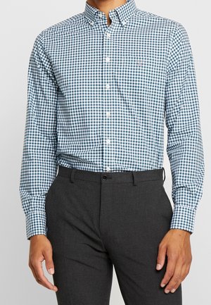 Uomo che indossa una camicia a quadri blu e bianca infilata in pantaloni eleganti grigio scuro, su uno sfondo semplice.