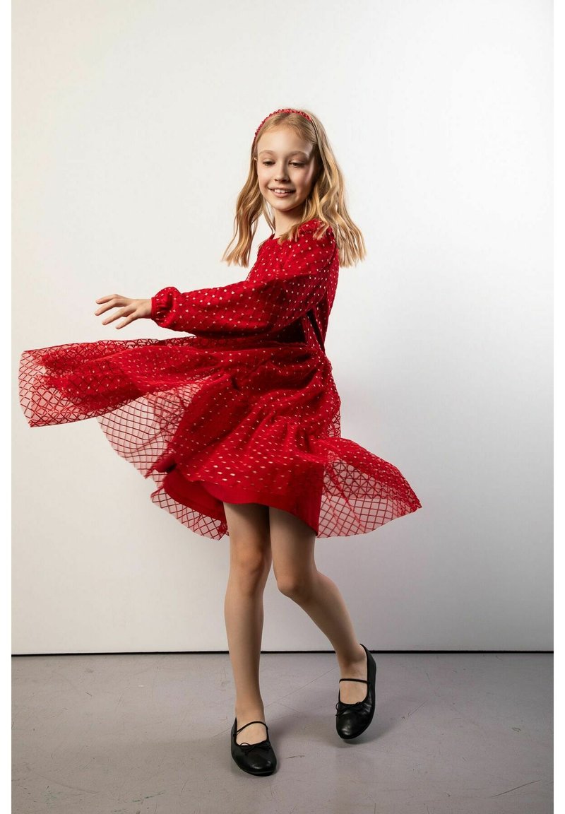 Czerwona sukienka z długimi rękawami, z siateczkową nakładką ozdobioną wzorem diamentowym oraz paskiem w talii. Czarne buty. Poza wirująca.