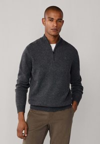 Gilet zippé gris foncé avec des poignets et un ourlet côtelés, fabriqué en matière tricotée douce, porté sur un T-shirt blanc, associé à un pantalon marron clair.