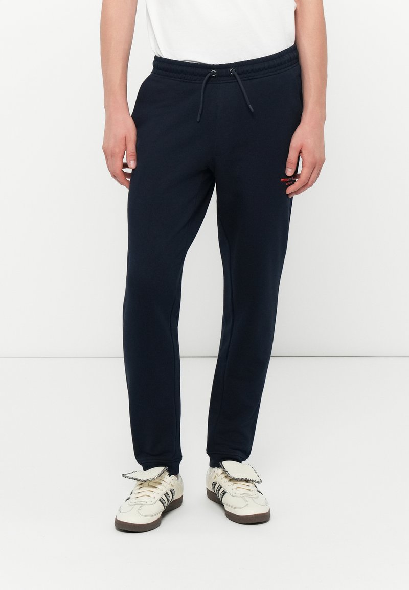 Mørkeblå joggingbukser med elastisk talje, snor og sidelommer, kombineret med beige sneakers med sorte detaljer og klapper.