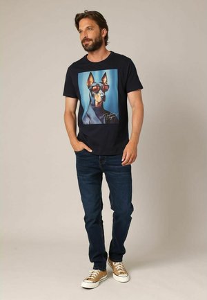 Camiseta navy con un gráfico de un perro usando gafas de sol. Combinada con jeans oscuros y zapatillas color beige. El modelo se encuentra de pie frente a un fondo neutro.