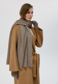 Sciarpa beige drappeggiata su un cappotto marrone chiaro. La sciarpa è realizzata in un tessuto morbido e presenta un'etichetta. Il cappotto ha maniche larghe e una vita legata.
