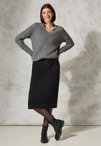 Grauer gerippter Strickpullover mit V-Ausschnitt, kombiniert mit einem schwarzen Midikleid mit Kordelzug und Fronttaschen. Schwarze Schnürstiefel.