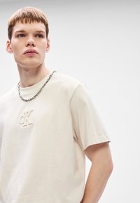 Ανοιχτό μπεζ βαμβακερό t-shirt με κεντημένο λογότυπο "cK", κοντά μανίκια και στρογγυλή λαιμόκοψη; συνδυασμένο με ασημένιο περιδέραιο.