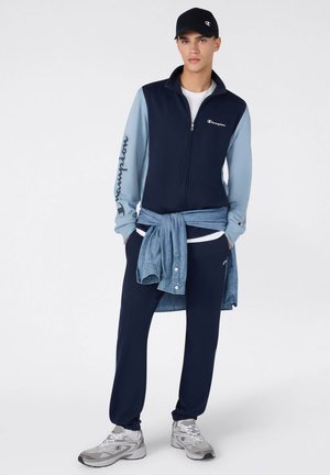 Tuta blu navy con maniche azzurre, con zip frontale; camicia di jeans a strati legata in vita; sneakers grigie. Logo Champion visibile.