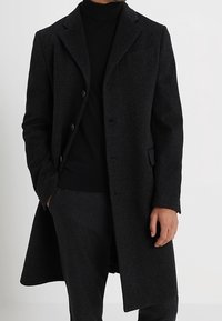 Cappotto lungo di lana nera con un design sartoriale, rever a punta e tre bottoni frontali. Presenta una tasca sul petto e tasche laterali.