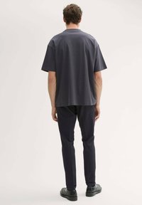 Graues Kurzarmshirt mit lockerer Passform, aus weichem Stoff. Getragen mit dunklen, taillierten Hosen und schwarzen flachen Schuhen, von hinten betrachtet.
