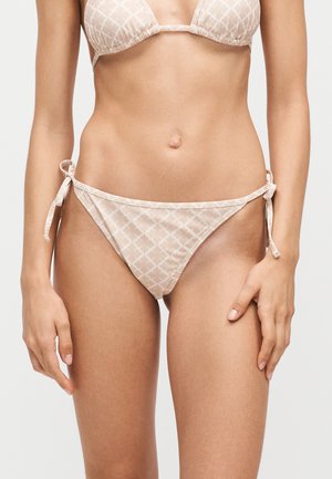 Kobieta nosząca jasnobeżowy dół od bikini z wiązaniami po bokach i subtelnym geometrycznym wzorem na jednolitym tle.