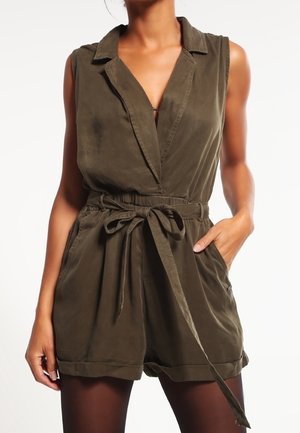 Sleeveless olive green romper with collar, deep V-neck, belted waist, and side pockets worn with black tights.