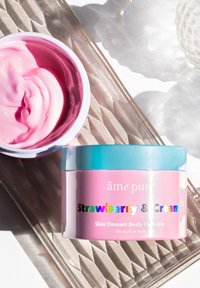 Rosa grädde i en rund burk med ett turkos lock. Etiketten säger "Jordgubb & Grädde" med iriserande bokstäver. Slät konsistens synlig inuti.