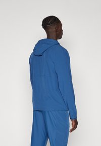 Modrá kapuce bunda s hladkým materiálem, elastickými manžetami a minimalistickým designem. Zobrazená ze zadního pohledu s jednoduchým siluetou.