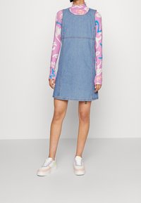 Denim ärmlös klänning med rak skärning och ljus tvätt, kombinerad med en mönstrad långärmad topp och pastellfärgade sneakers.