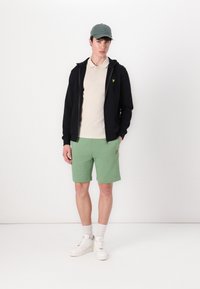 Hanorac negru cu fermoar, polo bej, pantaloni scurți verzi, adidași albi, șapcă gri. Logo brodat pe polo și pe pantaloni scurți. Material din amestec de bumbac.