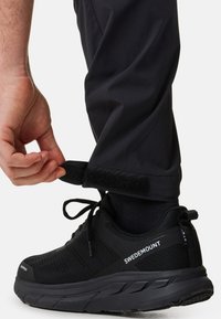 Black athletic shoe with textured upper, tied laces, and branding. Hand adjusting adjustable pant cuff with Velcro fastening.