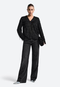 Juodas fringiniai topas su V formos iškirpte ir ilgomis rankovėmis, derinamas su juodais aukšto liemens plačiais džinsais ir sidabriniais smailiais batais. Laiko juodą karoliukų delninę.