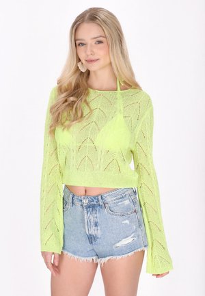 Ung kvinde med langt blondt hår iført en neongrøn hæklede sweater over en matchende top og slidte lyseblå denimshorts.