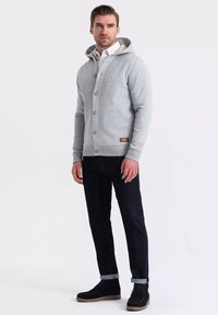 Felpa grigia in morbido tessuto con chiusura a bottoni, dotata di cappuccio e di un piccolo patch con logo. Abbinata a jeans scuri e scarpe nere.