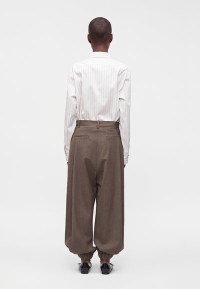 Personne vue de dos portant une chemise blanche à fines rayures rentrée dans un pantalon marron ample avec des poignets froncés et des chaussures noires sur un fond uni.