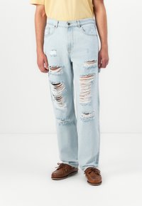 Light blue ripped jeans with multiple distressed patches and frayed edges, paired with brown boat shoes and a yellow shirt.