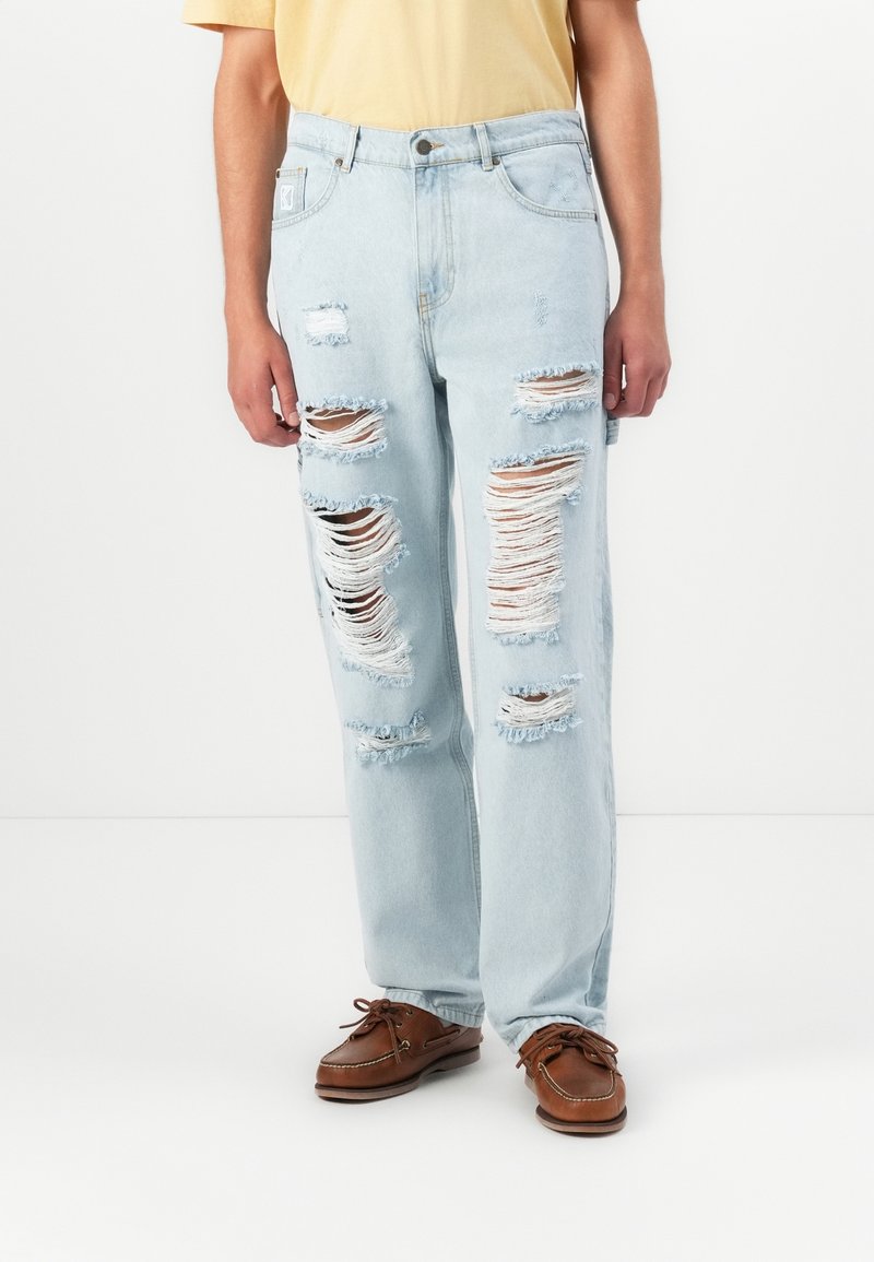 Light blue ripped jeans with multiple distressed patches and frayed edges, paired with brown boat shoes and a yellow shirt.