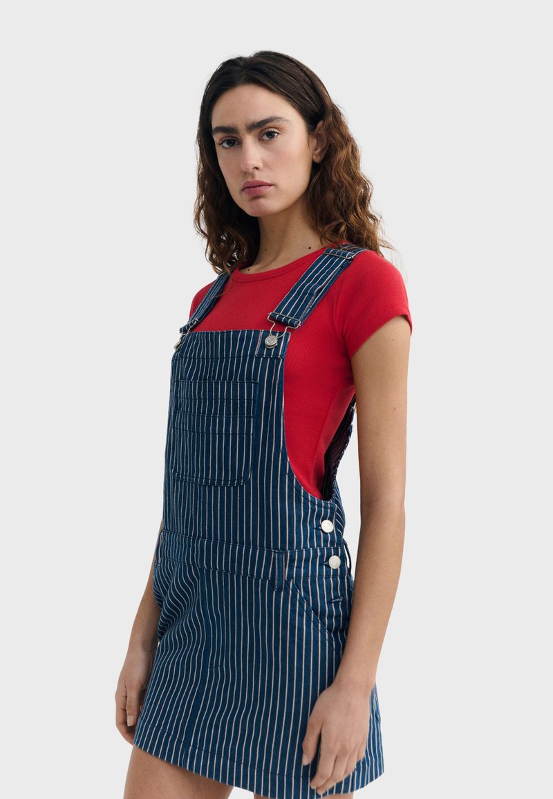 Femme aux cheveux ondulés portant une chemise rouge à manches courtes et une robe salopette bleue à rayures, se tenant devant un fond clair uni.