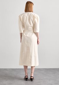 Rochie lungă, crem, cu mâneci până la trei sferturi și talie ajustată. Fabricată dintr-un material texturat, are un croi drept și o lungime până la jumătatea gambei.