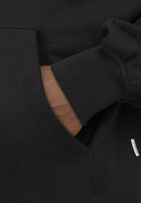 Zwarte hoodie met een kangoeroe-pocket, ribbelboorden en een zachte textuur. Een klein label is zichtbaar nabij de onderste zoom.
