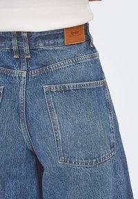 Jeans in denim con un lavaggio blu chiaro, dotati di un'etichetta in pelle marrone, due tasche posteriori e cuciture visibili lungo le cuciture.