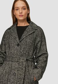Płaszcz w wzór houndstooth w czerni i bieli, z wyprofilowanym kołnierzem, pojedynczym zapięciem na guzik oraz przewiązywanym paskiem w talii. Fakturowany materiał.