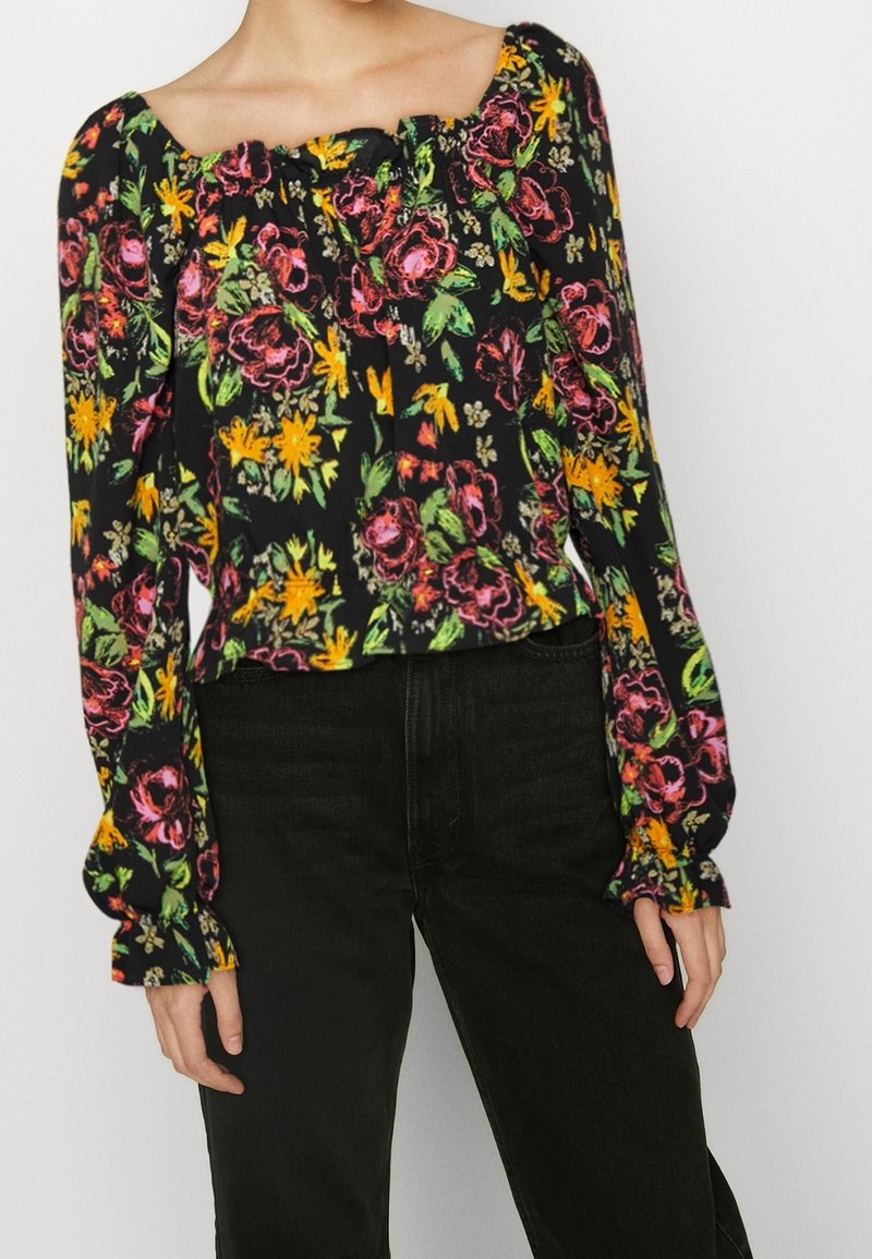 Zwarte bloes met bloemenpatroon, featuring levendige gele, roze en groene bloemen, vierkante halslijn, pofmouwen en geplooide manchetten. Gecombineerd met zwarte jeans.