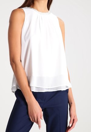Blouse blanche sans manches avec devant plissé et encolure ornée portée avec un pantalon bleu marine foncé sur une personne debout.