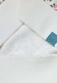 Baumwollstoff mit geripptem, strukturiertem weißen Kragen und floralem Muster; umfasst ein blaues Etikett mit einem Symbol. Weicher, weißer Stoff darunter.