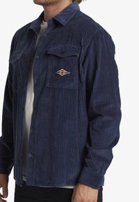 Navy corduroy shirt with vertical stripes, button-down front, two chest pockets, and a small embroidered logo on the left pocket.