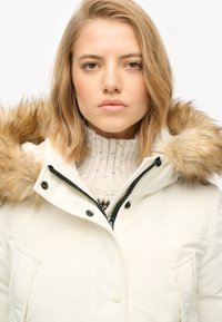 Hvid parka jakke med faux pels hætte, sort lynlås og trykknapper. Højhalset striktrøje i lyse beige, der titter frem under.