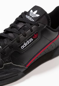 Czarne skórzane sneakersy z czerwonymi i niebieskimi pasami, posiadające czarny język z białym logo Adidasa oraz fakturowaną podeszwę. Sznurówki są czarne.